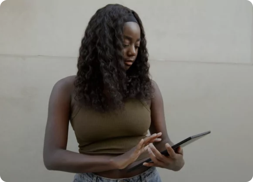A person with long curly hair, wearing a sleeveless top and jeans, is standing against a plain wall while looking at and tapping on a tablet.